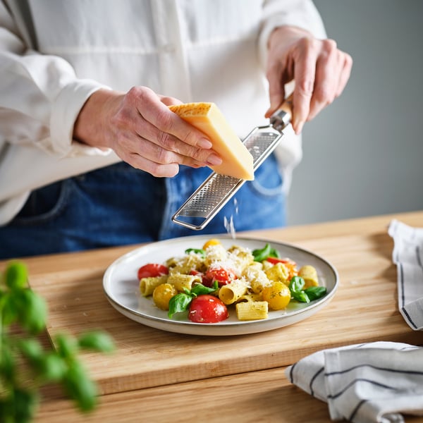 Grating cheese on pasta with tomatoes and basil using a metal zester with wooden handle.