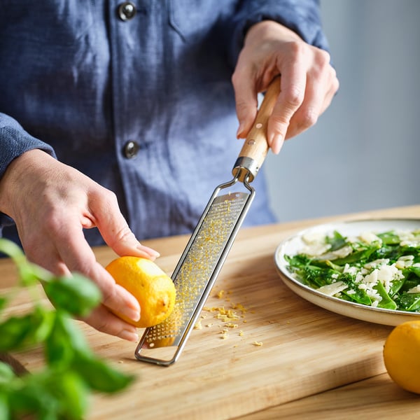 Person grates citrus zest using VARDAGEN zester on a wooden cutting board. Lemon zest collects in the grater.
