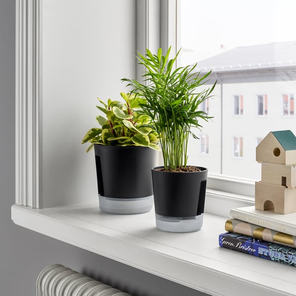 Two VÅRDTRÄD self-watering plant pots in black on a white shelf, surrounded by books and a wooden toy house.