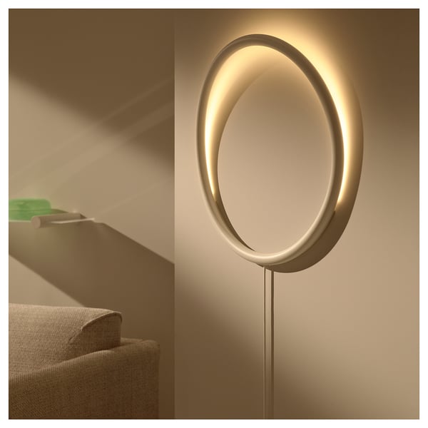 Modern oval led wall lamp VARMBLIXT emits soft light, mounted on beige wall beside beige sofa, sleek design.