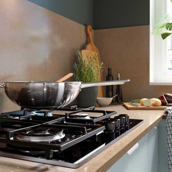 Kitchen scene: a gas stove with a black finish holds a wok. Nearby are herbs, cutting boards, and knives. The countertop is light-colored.