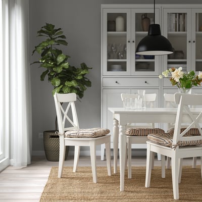 Cosy dining room with table, chairs, plant, glassware, flowers, cabinet, light, walls, curtains, rug.