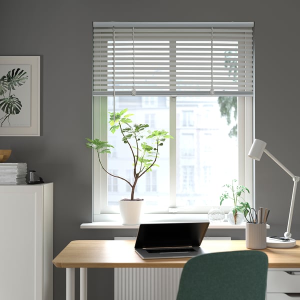 Modern home office with large VECKLARFLY blinds. Dark walls, wooden desk, green chair, and plants. Blinds partially closed, letting in natural light.