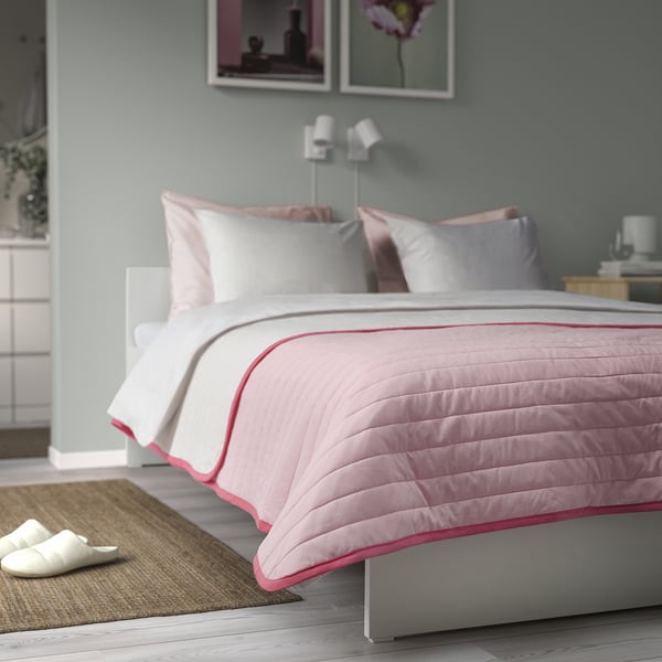 Neatly made bed with VETEJORDFLY pink/white bedspread, balanced pillows, and white slippers beside it.