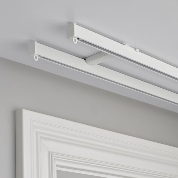 Two white ceiling brackets hold horizontal rails for curtain solution. Mounts with two screws. Creates space for smooth curtain glide.