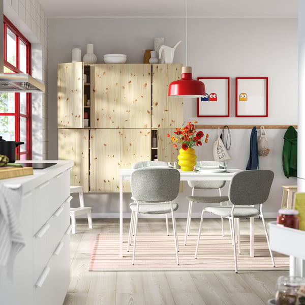 Modern kitchen with wooden cabinets, white dining table, colourful chairs, red pendant light, and yellow vase with flowers.