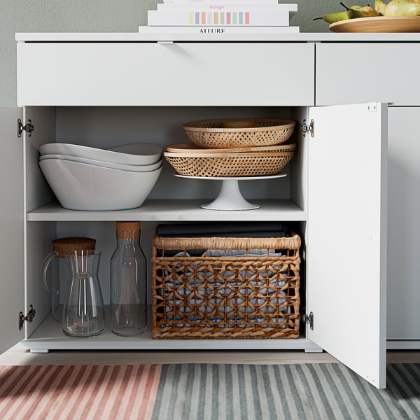 White cabinet with two shelves, bowls, trays, jars, and basket, grey rug below.