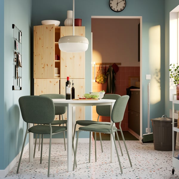 Small round white dining table with green chairs, set against light blue wall, with wooden cabinet and kitchen entrance nearby.