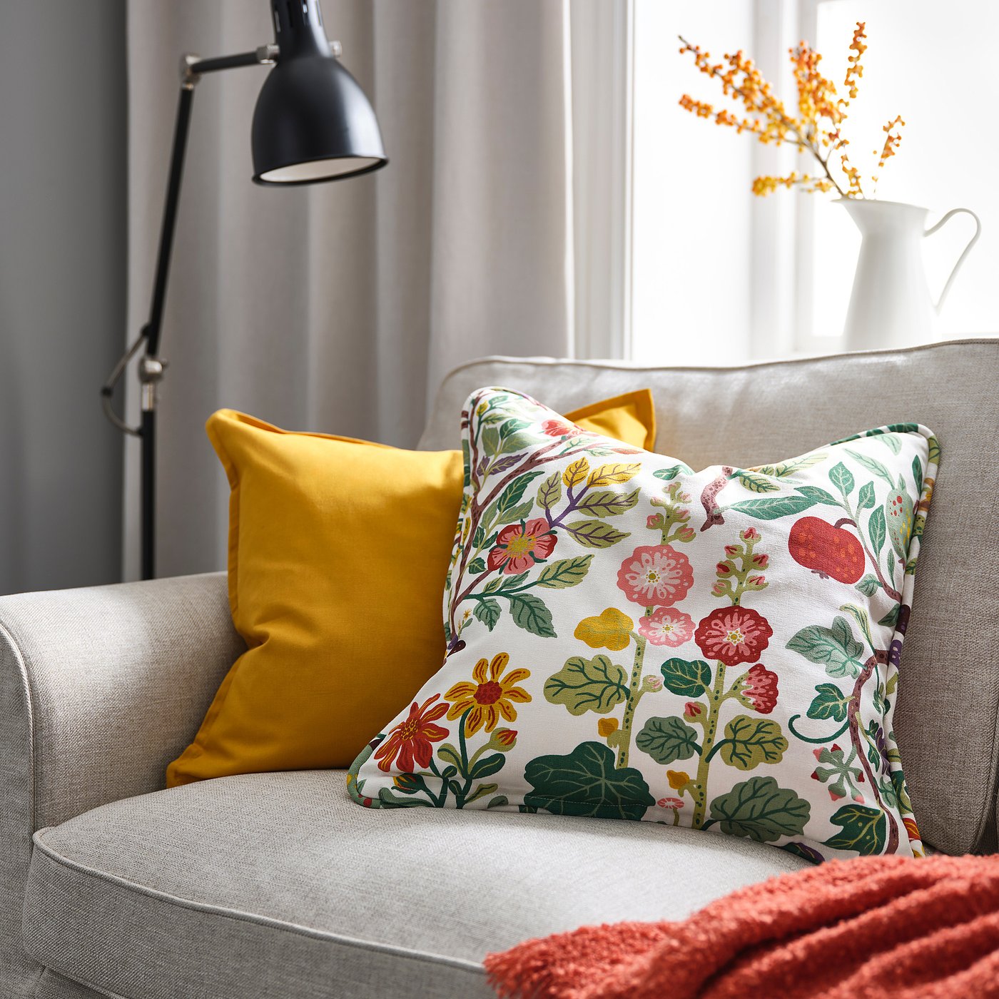 Cozy chair with VILDPERSILJA floral cushion, larger mustard pillow, red blanket, and orange flowers in a white vase.