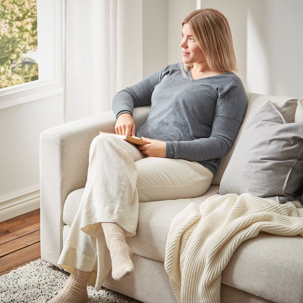 Person sitting on grey VIMLE sofa, using tablet, cosy setting with blankets, large window, light curtains.