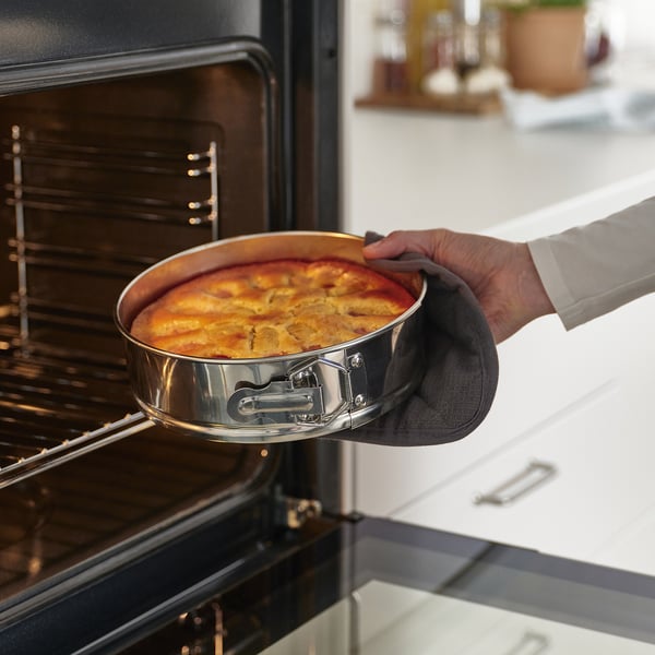 Person using oven mitts to pull out a golden-brown baked dish from an oven. The stainless steel springform pan ensures even heat distribution.