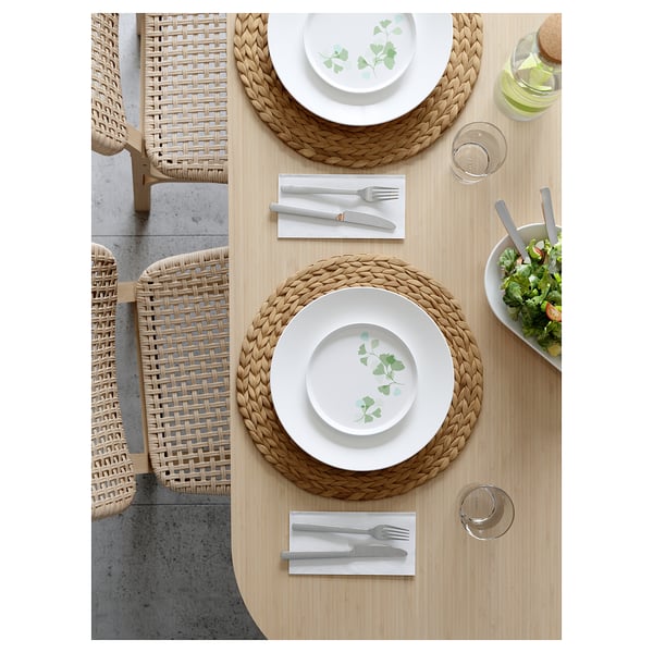 Minimalist dining setup with wicker chairs, placemats, white plates, silverware, green leaf pattern, salad bowl, and glassware on wooden table.