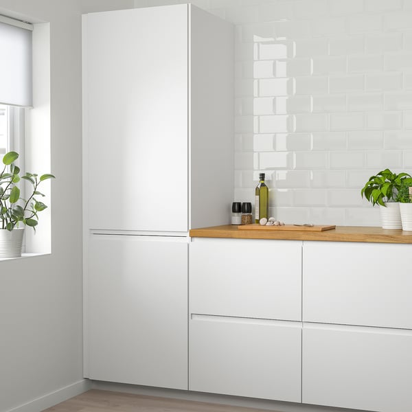 Modern white VOXTORP kitchen featuring tall refrigerator, sleek white cabinets, wood countertop, and potted plants.