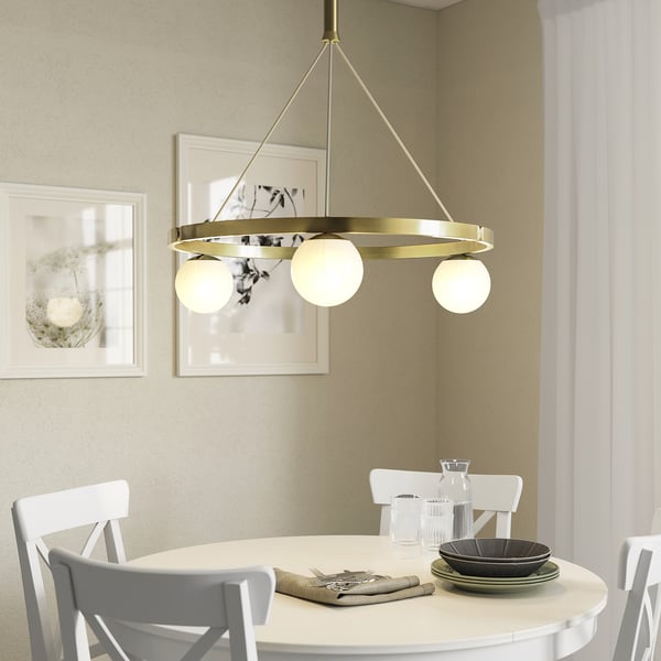 Misiņa piekaramā lampa ar trim stikla globusiem virs ēdamgalda, kas izstaro siltu gaismu.