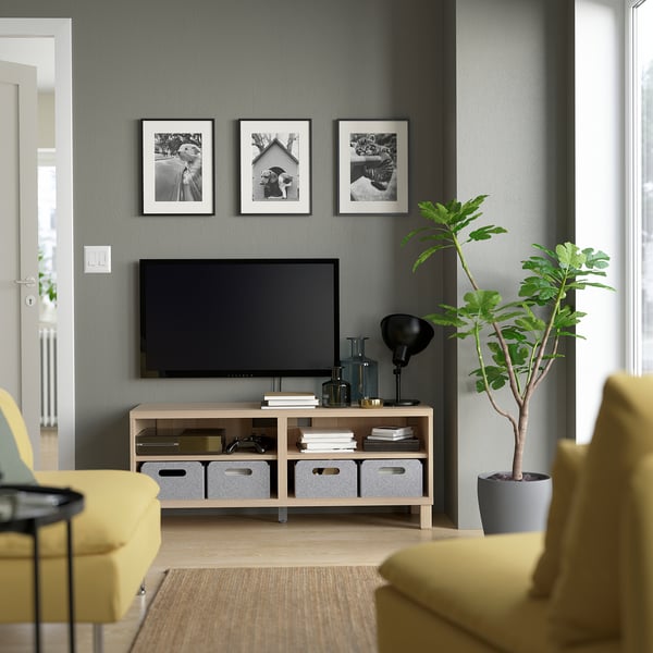Pelēka dzīvojamā istaba ar BESTÅ tv soli, plakanu ekrānu, dzelteniem krēsliem, zaļu augu, melnu lampu un fotogrāfijām virs televizora.