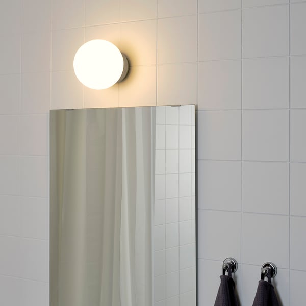 Apaļa, sienas lampa virs vannas istabas spoguļa, kas nodrošina mīkstu, vispārēju apgaismojumu. Melni dvieļi karājas uz metāla āķiem pa labi.