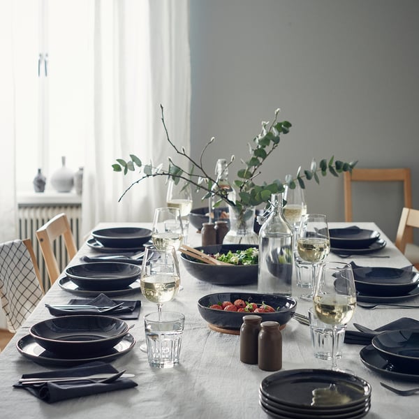 Eleganta ēdamistaba ar tumši pelēkiem GLADELIG dziļiem šķīvjiem, caurspīdīgām glāzēm, kārtīgi salocītām salvetēm un centrālo vāzi ar zaļumiem.