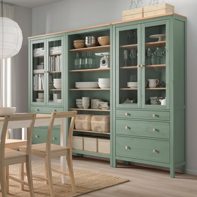 Zaļā HEMNES glabātuve, stikla durvis, koka plaukti, bļodas, trauki, vīna glāzes, grāmatas, krēsli.