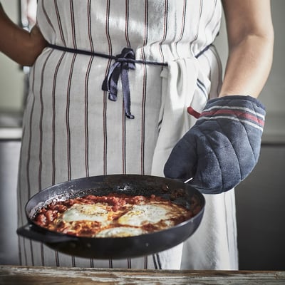 Cilvēks pelēkā svītrainā priekšautā tur pannu ar shakshuka, izmantojot melnu cepeškrāsns cimdu.