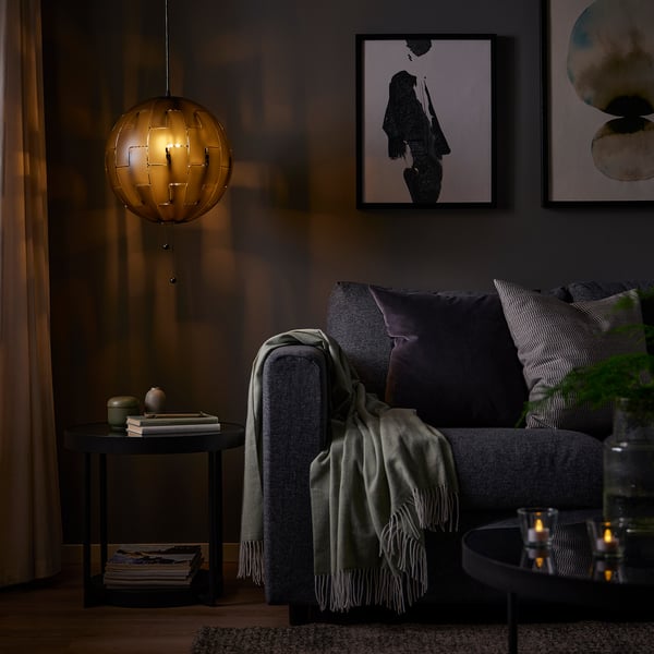 Mājīga dzīvojamā istaba ar IKEA PS 2014 lampu virs tumša dīvāna, kas raida siltu gaismu. Sānu galdiņā ir grāmatas un dekori, māksla uz sienas.