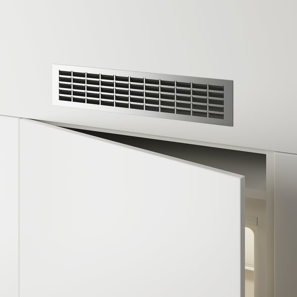 Taisnstūrveida metāla ventilācijas režģis, kas uzstādīts uz baltas sienas virs durvīm, kas nodrošina gaisa plūsmu.