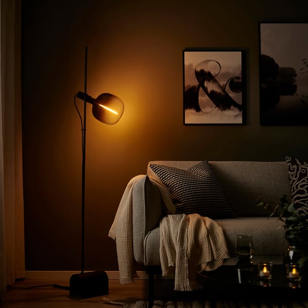 Mājīga dzīvojamā istaba ar melnu grīdas lampu, siltu gaismu, pelēku dīvānu, spilveniem, segām un sienas mākslu. Lampa ir augsta un plāna ar unikālu spuldzi.