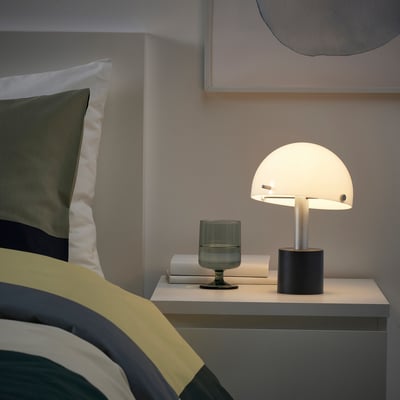 Mūsdienīga guļamistaba ar NÖDMAST lampu uz nakts; Lampa ir maza, balta un kupola formas, blakus zaļai glāzei un grāmatai.