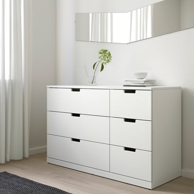 Ikea NORDLI 6 atvilktņu kumode pie sienas, minimālisma istaba, vāze ar zaļu ziedu, grāmatas.