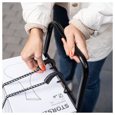 Persona stumj melnus RULLEBÖR ratiņus, pārvadājot baltu kasti ar marķējumu sto & s. Viņi valkā baltu džemperi un smilškrāsas aproces.