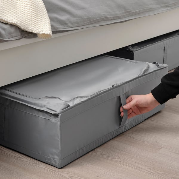 Cilvēks, kas velk pelēku SKUBB glabāšanas kasti zem gultas, ideāli piemērota sezonas priekšmetu glabāšanai, aizsardzībai no putekļiem un vietas taupīšanai.