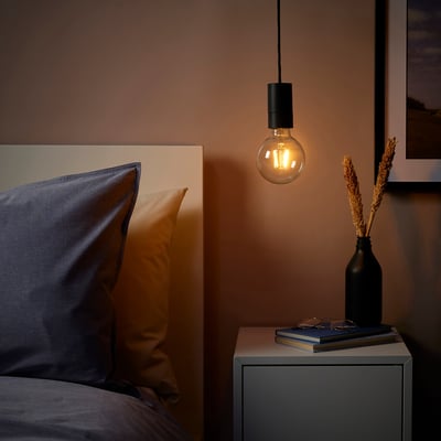 SUNNEBY piekaramā lampa ar melnu auklu, silta spuldze blakus gultai ar spilveniem. Naktsskapī ir ziedi un grāmatas, mājīga atmosfēra.