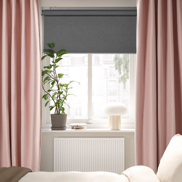 Guļamistabas logs ar pelēku rullo žalūziju, mīksti rozā lineārās tekstūras aizkariem, baltu lampu un augstu zaļu augu uz palodzes, baltu radiatoru zemāk.