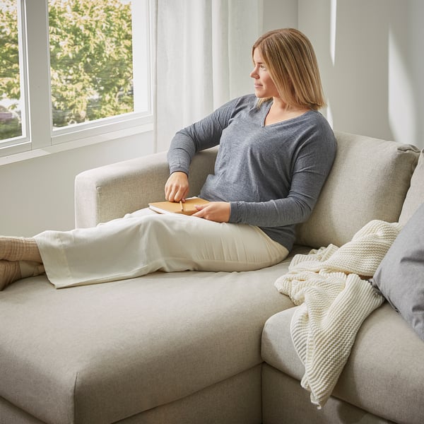 Sieviete atpūšas pelēkā VIMLE dīvānā pie liela loga, ar gaišām segām un spilveniem.