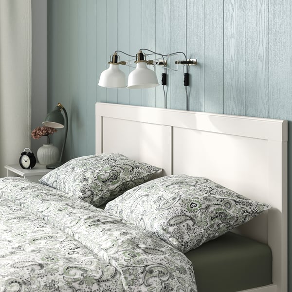 Уютная сцена в спальне с белой деревянной кроватью, узорчатыми простынями, двумя настенными лампами над кроватью и прикроватной тумбочкой с лампой и часами.