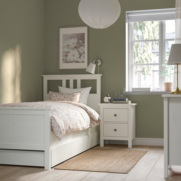 Уютная спальня с белой кроватью HEMNES, стенами из шалфея, постельным бельем с цветочным рисунком, тумбочкой с лампой, книгами и окном с естественным освещением.