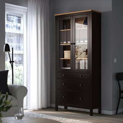 Черная деревянная витрина HEMNES со стеклянными дверцами и ящиками стоит рядом с окном и стулом.