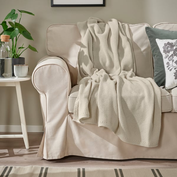 Бежевый диван с мягким вязаным не совсем белым пледом, накинутым на него. На небольшом столике рядом с ним стоит ваза с растением.