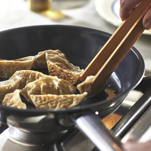 Рукой палочками для еды можно выбрать пельмени из черной сковороды на плите, подчеркивая антипригарную поверхность сковороды и простоту приготовления.