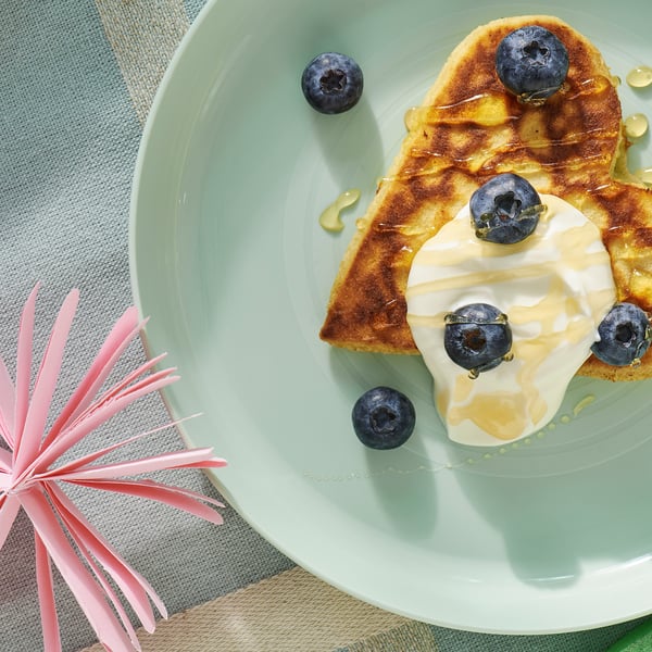 На светло-зеленой детской тарелке стоит вкусный черничный блинчик, покрытый йогуртом и сбрызнутый медом, и все это на уютной голубой скатерти.