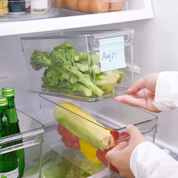 Человек раскладывает продукты в холодильнике с помощью прозрачных, штабелируемых ящиков для хранения. Под ручкой закреплена записка с датой.