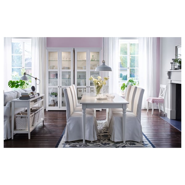 Элегантная столовая с белым столом, стульями и шкафом. Большие окна со шторами, растениями и стильным подвесным светильником.