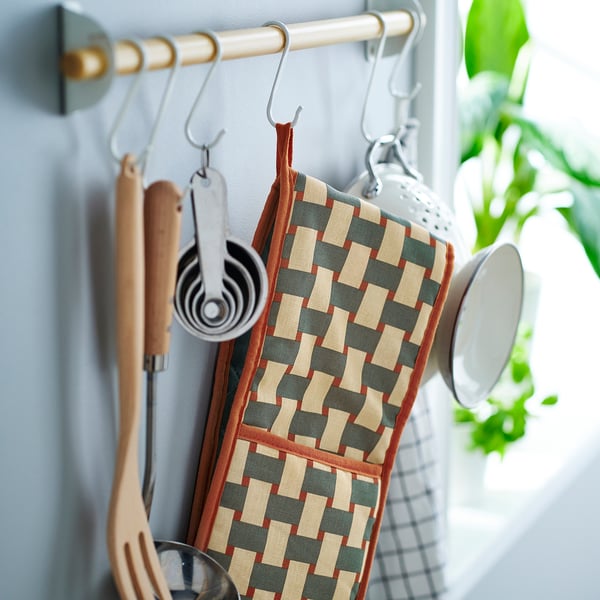 Кухонная сцена с клетчатой прихваткой на крючке, рядом с посудой и кружками, подчеркивающая ее безопасную двойную конструкцию.