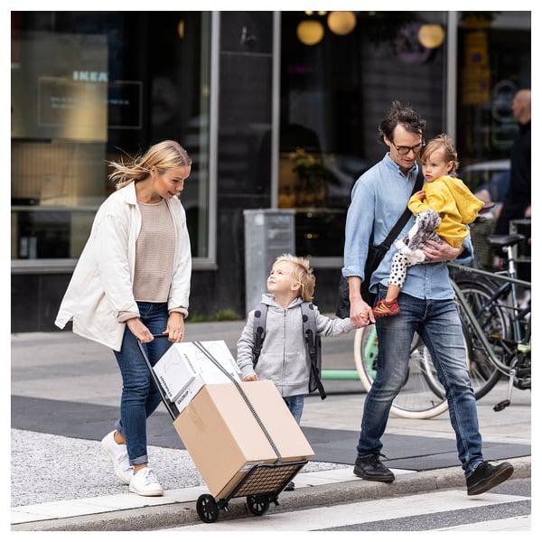 Семья несет бежевую тележку для покупок IKEA по улице. Папа держит ребенка и плюшевую игрушку, мама тянет тележку с еще одним ребенком рядом.