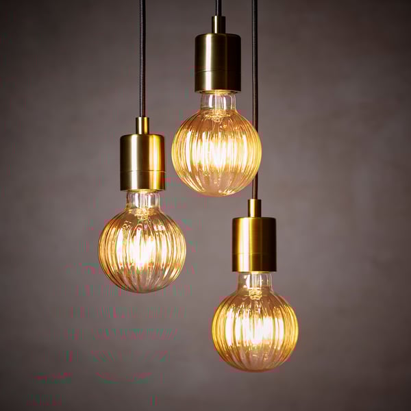 Три современных подвесных светильника с ребристыми стеклянными колбами свисают на шнурах золотистого цвета. Лампочки подсвечиваются, отбрасывая теплое свечение.
