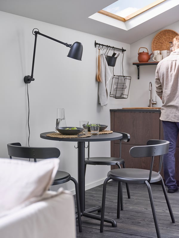 Современный кухонный уголок с черным круглым столом STENSELE, тремя стульями, настенным светильником и человеком, готовящим еду.