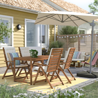 Патио на заднем дворе с желтым домиком, деревянным столом, четырьмя стульями, бежевыми подушками, бежевым зонтом, полосатым ковром и забором для уединения.