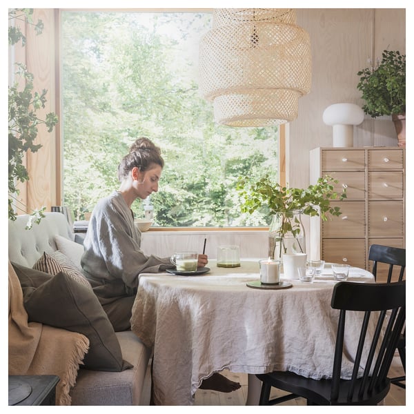 A person enjoys a meal by a large window in a cosy, natural-lit dining area with a beige tablecloth, wooden furniture, and green plants.