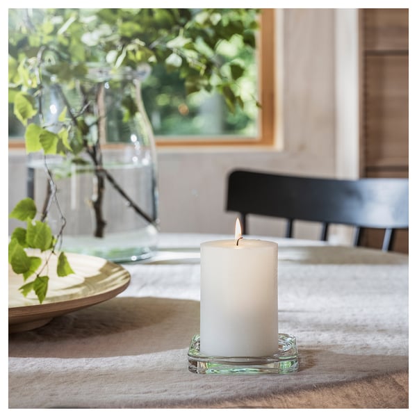 Cosy table setting: white lit candle in glass, surrounded by plates and vase with greenery.