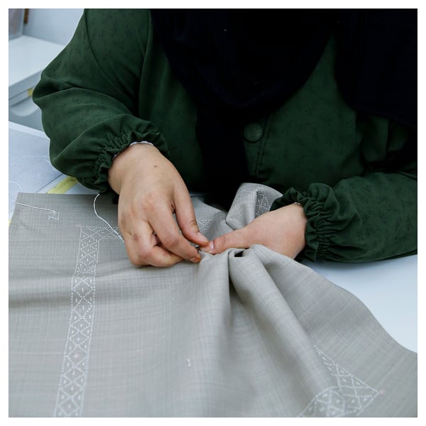 Person wearing green garment, sewing grey embroidered fabric by hand; intricate design and craftsmanship.