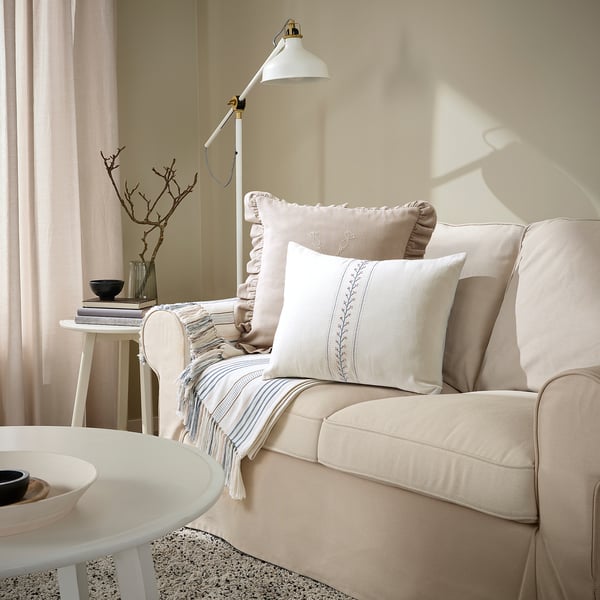 Cosy living room with beige sofa, ÅKERNEJLIKA cushion, decorative pillows, side table, and floor lamp.